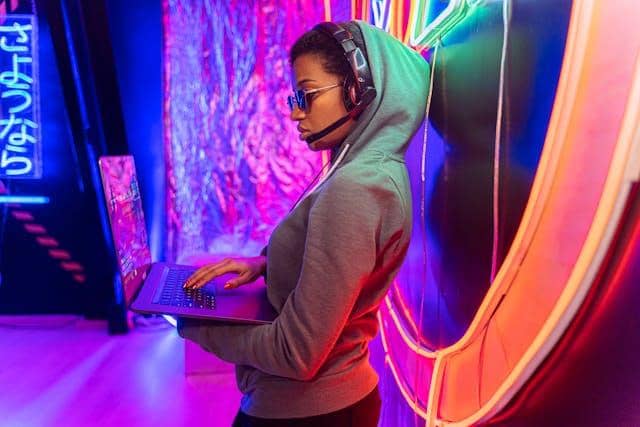 Cómo encontrar una laptop gamer que sí cumpla tus expectativas 24 Una mujer con auriculares y gafas de sol usa una laptop gamer en un espacio con iluminación neón.