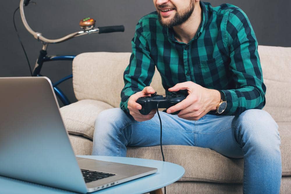 Cómo encontrar una laptop gamer que sí cumpla tus expectativas 22 Un hombre relajado juega videojuegos con un mando en su sofá, con un portátil a su lado.